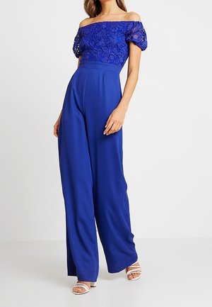 Donna che indossa una tuta blu royal con scollo a barca, dettagli in pizzo sulla parte superiore e pantaloni a gamba larga, abbinata a tacchi bianchi con punta aperta.