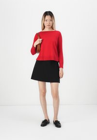 Červený pletený sveter s okrúhlym výstrihom, skombinovaný s čiernou sukňou do áčka. Modelka nosí čierne topánky a drží hnedú kabelku.