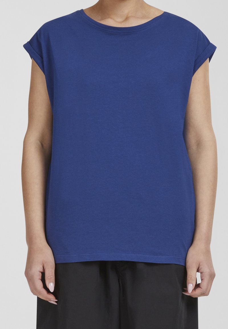 Personne portant un T-shirt bleu uni à manches courtes retroussées et un pantalon noir, debout les bras détendus le long du corps.