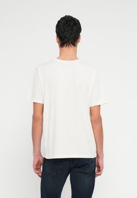 Biała bawełniana koszulka z krótkimi rękawami, okrągłym dekoltem i luźnym krojem, pokazana od tyłu na jednolitym białym tle.