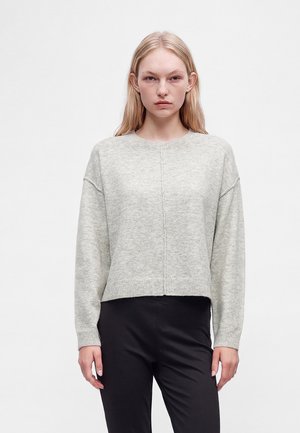 Femme blonde portant un pull en maille gris clair à manches longues et un pantalon noir, debout devant un fond blanc uni.