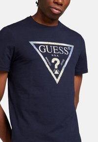Camiseta de algodón azul marino con un gráfico triangular que presenta el texto "GUESS", un signo de interrogación y acentos desgastados. Manga corta, cuello redondo.