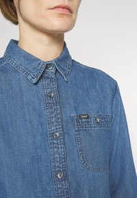 Denimskjorta med klassisk krage, knappstängning fram och en bröstficka med en logotagg. Tyget är blått med en strukturerad yta.