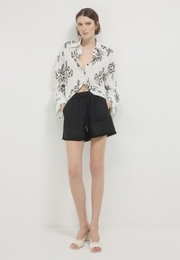 Camicia bianca con bottoni e motivi astratti neri, abbinata a pantaloni corti neri con coulisse, dotati di due tasche e orli sfrangiati. Mule beige con punta aperta.