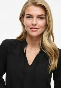 Melnas blūzes ar V formas izgriezumu, garām piedurknēm un frāzētiem plecu detaļām. Gluda tekstūra ar pakarināmo saitēm izgriezumā.