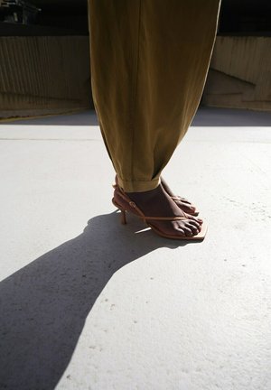 Pies de piel oscura con sandalias marrones de tacón alto con tiras, vistiendo pantalones verde oliva, proyectando una sombra larga sobre un suelo de hormigón claro.