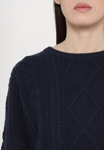 Pull en maille bleu marine avec un motif texturé comprenant des torsades et des losanges. Col côtelé. Tissu doux et épais.