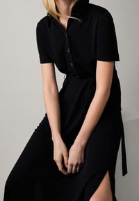 Sort skjortekjole med krave, korte ærmer og en tætsiddende pasform. Har en sideslids og en bæltestropp i taljen for form og struktur.