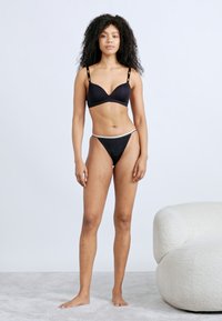 Musta alusasusetti, joka sisältää säädettävin olkaimin varustetun push-up-rintaliivit ja matalavyötäröisen stringit, esillä mallin päällä vaaleaa taustaa vasten.