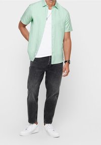 Uomo che indossa una camicia a maniche corte verde menta con bottoni sopra una maglietta bianca, jeans neri, sneakers bianche e un orologio da polso, in piedi contro uno sfondo chiaro.