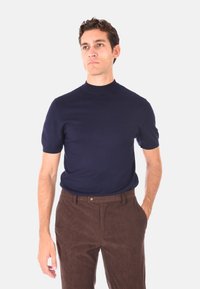 Uomo che indossa un maglione a maniche corte blu navy con colletto alto e pantaloni in velluto a coste marroni, in piedi con una mano in tasca su sfondo bianco.