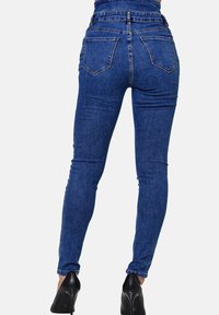 Vaqueros skinny de tiro alto en denim azul con un diseño ajustado, cinco bolsillos y un ligero desvanecimiento. Se llevan con zapatos de tacón negros.