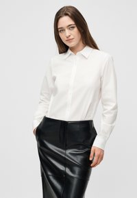 Νέα γυναίκα με καστανά μαλλιά φοράει λευκό πουκάμισο με κουμπιά, το οποίο είναι περασμένο μέσα σε μαύρη δερμάτινη φούστα, με τα χέρια στις τσέπες.