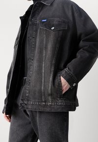 Chaqueta de mezclilla negra con cierre de botones al frente, bolsillo en el pecho y bolsillos laterales. Presenta costuras en contraste y una pequeña etiqueta con logo azul.