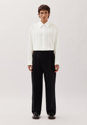 Giovane uomo che indossa una camicia bianca a righe verticali, pantaloni neri a gamba larga e scarpe eleganti nere, in piedi davanti a uno sfondo semplice.