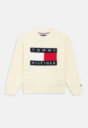 Crème ribgebreide trui met een opvallend rechthoekig logo waarop "TOMMY" en "HILFIGER" staan in zwart met rode accenten in het midden.