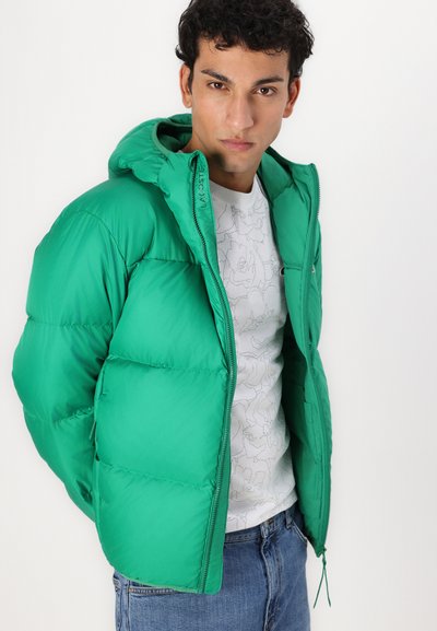 Veste coupe-vent verte avec capuche, fermeture éclair à l'avant et logo brodé. Portée par-dessus une chemise blanche à motifs et un jean bleu. Texture lisse.