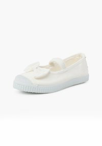 Zapatilla de tela blanca slip-on con puntera redonda, acento de lazo en la parte delantera y suela de goma texturizada de color azul claro.