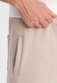 Beige Hosen aus strukturiertem Stoff mit Seitentaschen und sichtbaren Nähten. Eine Hand greift sanft den Bund.