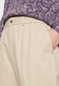 Béžové kalhoty s elastickým pasem a bočními kapsami, se stříbrným knoflíkem. Tkanina vypadá hladce. Vrchní díl má fialový vzor.