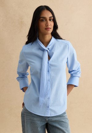 Vrouw met lang donker haar die een lichtblauw gestructureerd overhemd met knopen en een gebonden neksjaaltje en blauwe spijkerbroek draagt, staand met de handen achter haar rug.