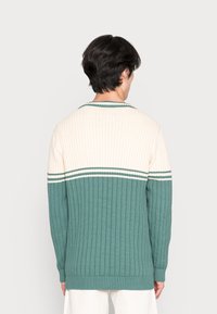 Jersey de punto acanalado con la mitad inferior verde y la mitad superior crema. Presenta rayas blancas contrastantes a lo largo de los hombros y en el dobladillo.