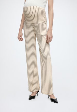 Gravid person iført beige graviditetsbukser med sorte lodrette striber og sorte spidse hæle, stående mod en ensfarvet baggrund.