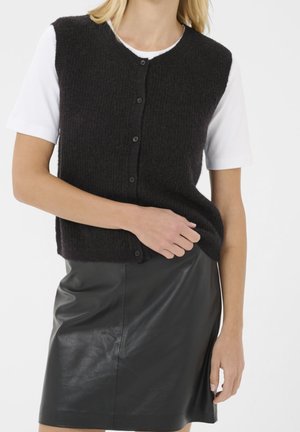Femme portant un gilet en maille noir boutonné sur une chemise blanche à manches courtes et une jupe en cuir noire, debout avec une main sur le gilet.