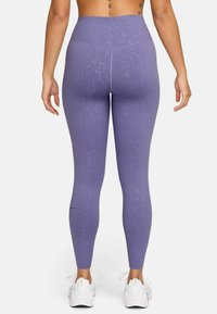 Hoch geschnittene, lila Leggings mit floralem Muster, aus dehnbarem Stoff, mit einem Logo nahe dem Knöchel und glatter Textur.