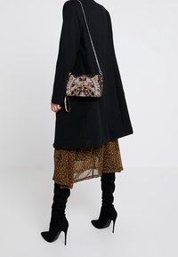 Cappotto nero con una gonna floreale marrone fluida e stivali neri al ginocchio. La persona porta una borsa decorativa con motivi intricati.