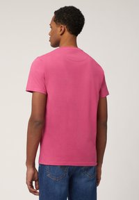 Camiseta de manga corta y cuello redondo en algodón de color rosa vibrante. Textura suave y corte relajado. Lleva puestas jeans de mezclilla azules, vista desde atrás.