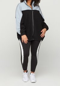 Personne portant une veste zippée noire et grise, des leggings noirs avec des bandes latérales blanches et des baskets blanches, se tenant contre un fond uni.