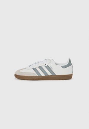 Vita sneakers med tre grå ränder, mocka tåhuvud, grå häldetalj och gummiyttersula. Har snörning och vadderad krage.