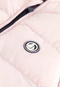 Šviesiai rožinė pūkinė striukė su lygiu paviršiumi, kurioje yra juoda užtrauktukas ir apvalus logotipo pleistras kontrastingų spalvų ant krūtinės.
