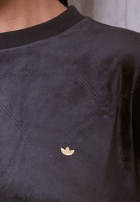 Detailní záběr na tmavě šedou texturovanou košili s vyšívanými diamantovými vzory a malým zlatým logem Adidas trefoil poblíž hrudníku.