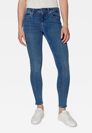 Person stående iført blå skinny jeans, hvide sneakers og en hvid skjorte, der er stukket ned i bukserne, vist fra skuldre til fødder mod en ensfarvet baggrund.