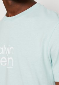 Camiseta de algodón de color azul claro con cuello redondo, que presenta un gran logo blanco de Calvin Klein en el frente y mangas cortas.