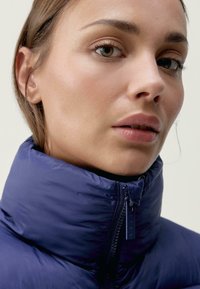 Temno modra izolirana jakna z visokim ovratnikom in zadrgo. Gladka tekstura z sijajem, zasnovana za toploto in udobje.