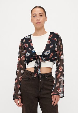 Musta läpinäkyvä crop-top, jossa on värikkäitä simpukkakuvioita, pitkät hihat ja edessä solmittava kiinnitys. Päällä valkoinen tiukka paita.