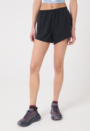 Femme portant un short de sport noir et des chaussures de course violettes, debout devant un fond blanc uni.