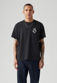 Sort bomulds t-shirt med korte ærmer og rund hals. Har et hvidt grafisk design på venstre brystområde. Bæres med mørkeblå jeans.