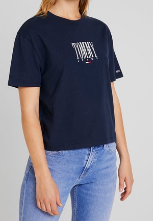 Personne portant un t-shirt Tommy Jeans bleu marine et un jean bleu clair, debout devant un fond gris uni.