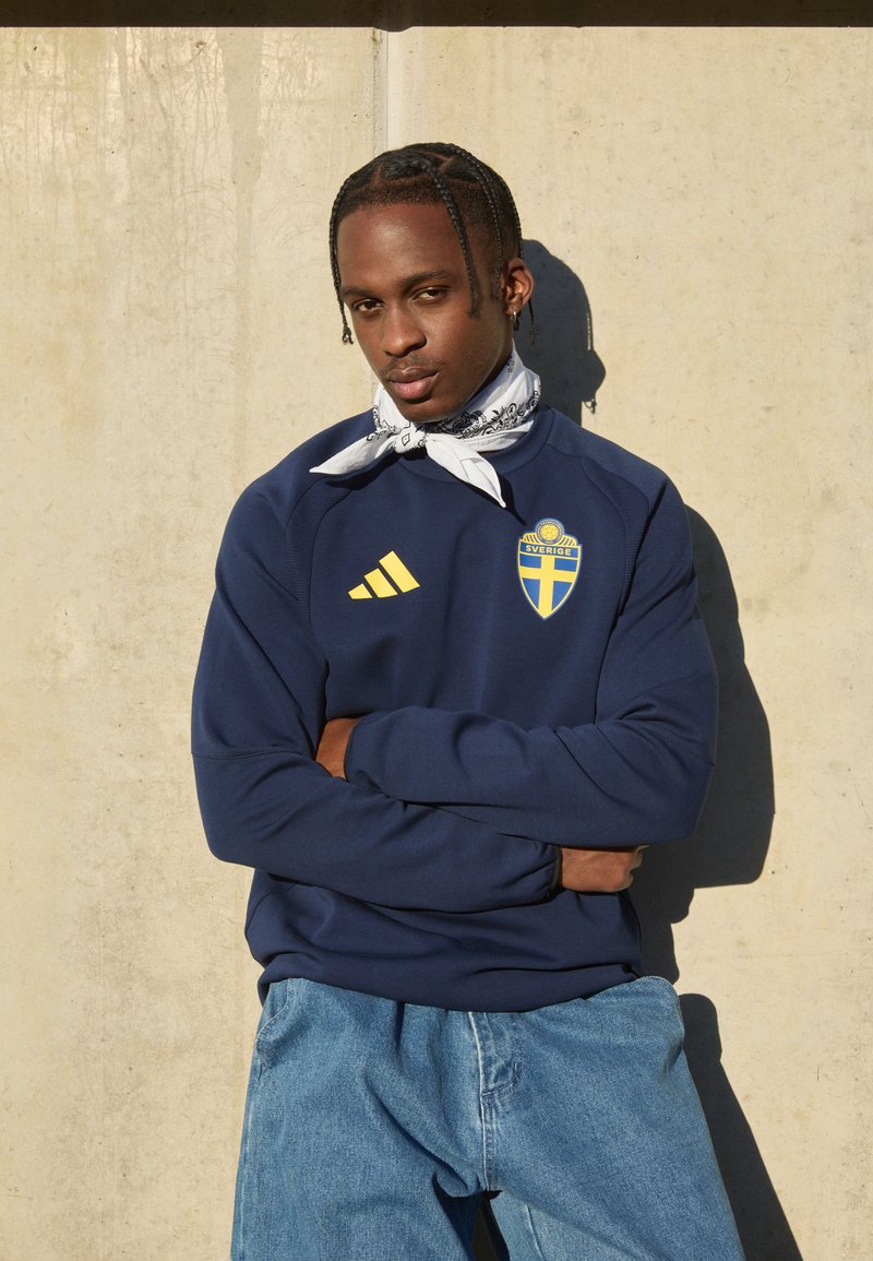 Mornarska sweatshirt s rumenim Adidas logotipom in grbom Švedske, kombinirana z svetlo modrimi kavbojkami. Bele šal okoli vratu dodaja kontrast. Gladka tekstura.