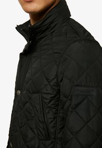 Giubbotto trapuntato nero con colletto alto, bottoni frontali e tasche laterali. Presenta un tessuto strutturato e un'etichetta con il marchio sul maniche.