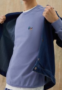 Felpa viola chiaro con scollatura a girocollo, presenta un piccolo logo multicolore. Sovrapposta a una giacca in denim scuro. Tessuto morbido.