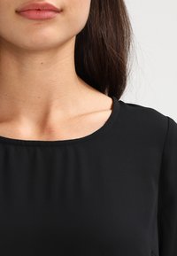 Haut noir avec un col rond, un tissu lisse et des détails de couture subtils le long des bords. Design minimaliste sans motifs.