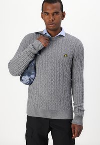 Suéter gris de punto trenzado con bordes acanalados y un pequeño logo amarillo. Usado sobre una camisa azul clara, combinado con pantalones oscuros y una bolsa estampada.