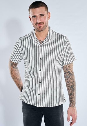 Mann mit kurzen Haaren und Tätowierungen, der ein schwarz-weiß gestreiftes Hemd mit kurzen Ärmeln und schwarzen Hosen vor einfarbigem Hintergrund trägt.