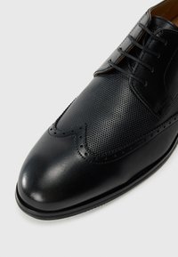 Chaussure habillée en cuir noir avec un motif texturé sur le dessus, des détails de brogue et des lacets noirs sur un fond blanc.