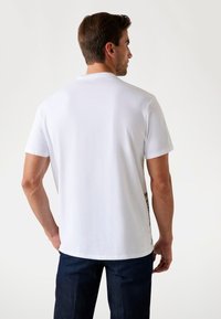 Hvid kortærmet T-shirt i bomuldsstof, rund halsudskæring, set fra ryggen, med et diskret design på den nederste side, parret med mørkeblå jeans.